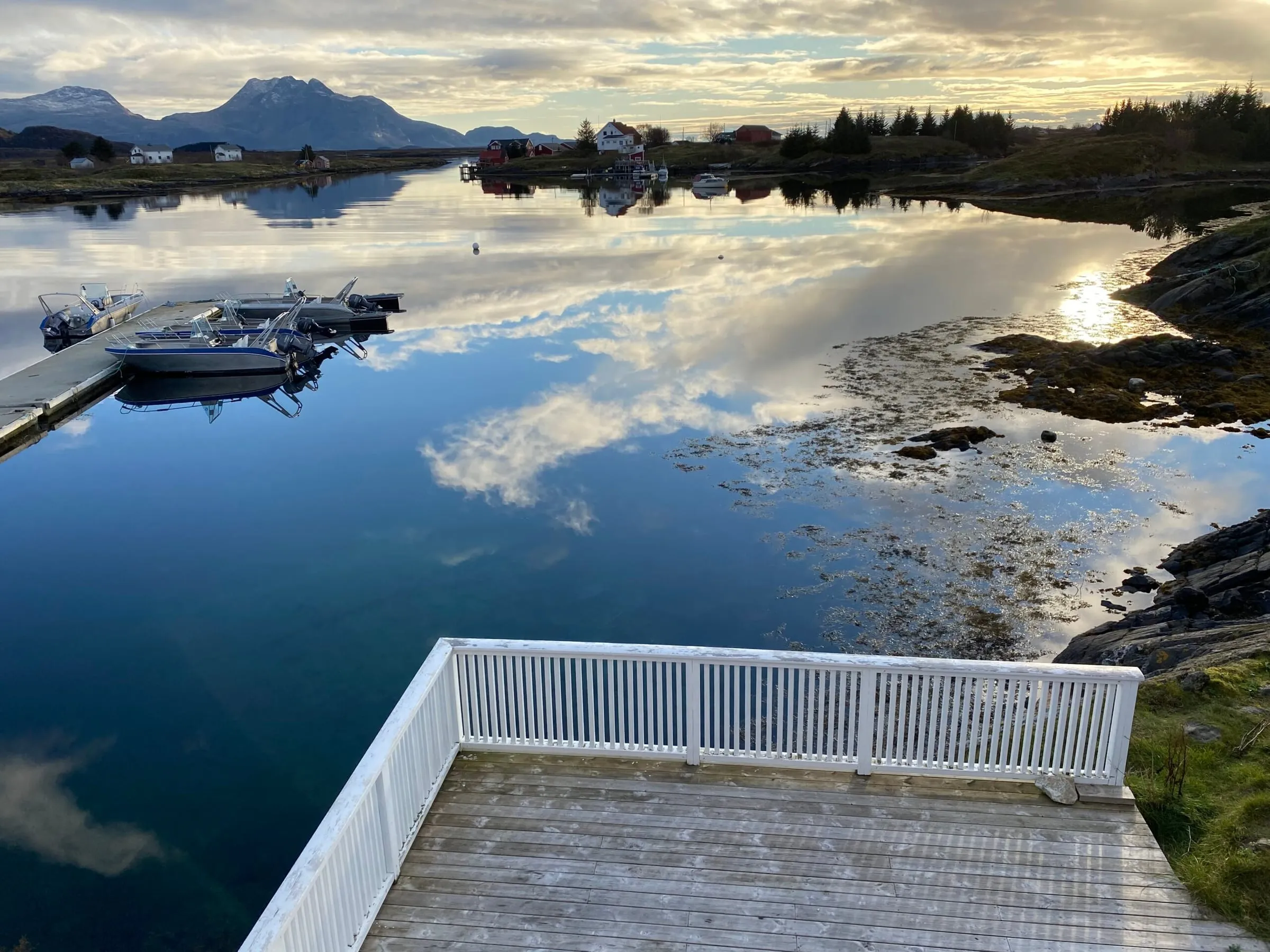 Blank sjø med speilende skyer, brygge og båter i havnen